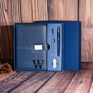 Puede incluir: Un juego de regalo azul que incluye un diario de cuero, un bolígrafo, un lápiz óptico y una goma de borrar. El diario lleva el nombre "William" y las iniciales "W" y "VV". El conjunto se presenta en una caja azul, sobre una superficie de madera.