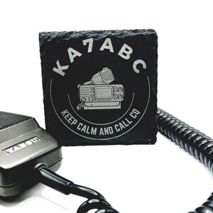 May include: Black square coaster with the text "KA7ABC" and a radio graphic. The coaster also has the text "KEEP CALM AND CALL CO". A black microphone and coiled cord are in the foreground.