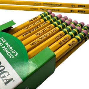 May include: A green cardboard box containing yellow Dixon Ticonderoga pencils with pink erasers and green metal ferrules. Each pencil is personalized with black text including names like Jackson, Lucas, Riley, and phrases such as Math Wizard, Homework Is Lame, You Got This, Mrs Cooper's Class, and Be Kind.