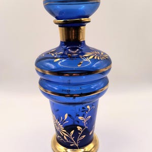 May include: A cobalt blue glass decanter featuring a wide, flared rim, a rounded body with two horizontal ridges, and a circular base. The glass is decorated with gold-painted floral patterns and horizontal gold bands around the neck and rim.