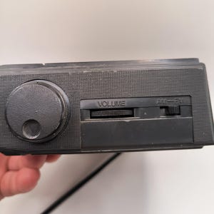 May include: A black vintage radio featuring a large volume knob and an AM/FM slider. The radio has a textured surface and a power cord. The word "VOLUME" is printed on the radio.