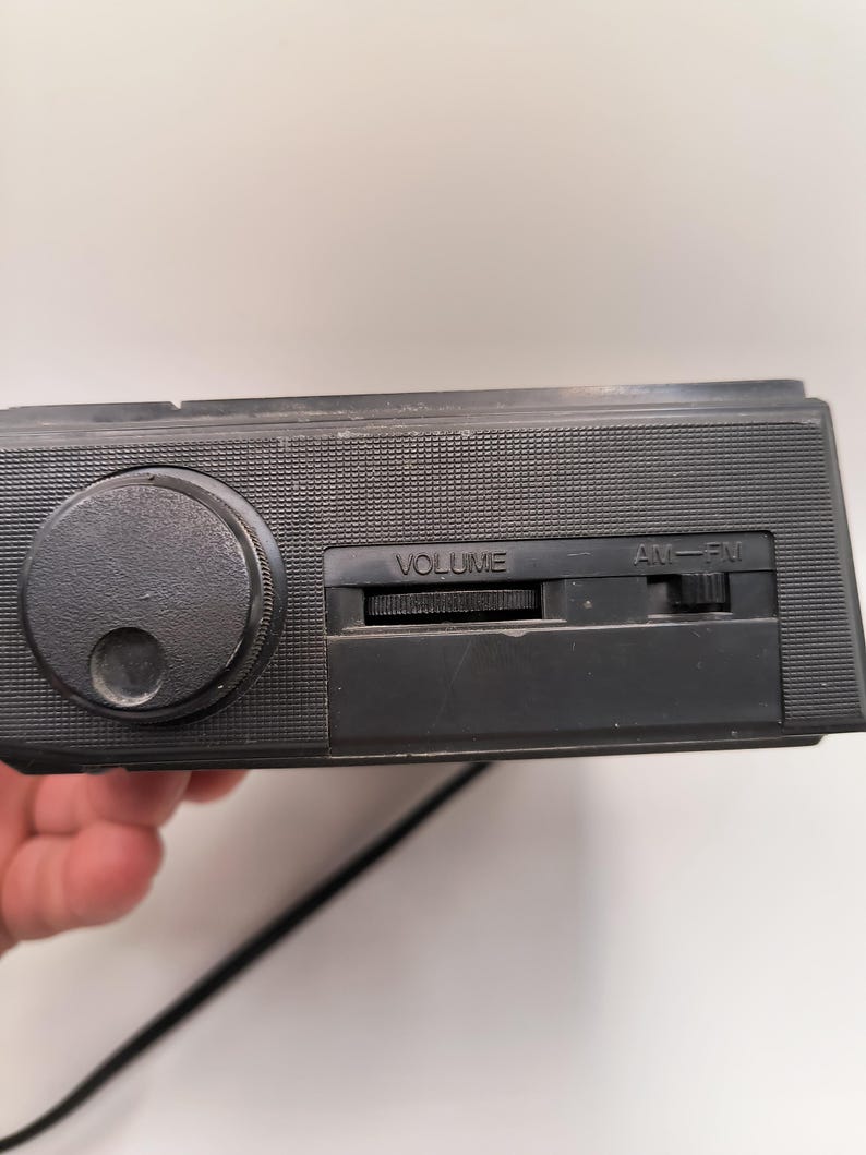 May include: A black vintage radio featuring a large volume knob and an AM/FM slider. The radio has a textured surface and a power cord. The word "VOLUME" is printed on the radio.