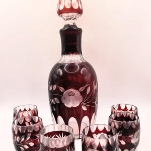 May include: A vintage, ruby-colored glass decanter set. The set includes a tall decanter with a stopper and six matching glasses. The decanter and glasses feature etched floral designs. The set is displayed on a white surface.