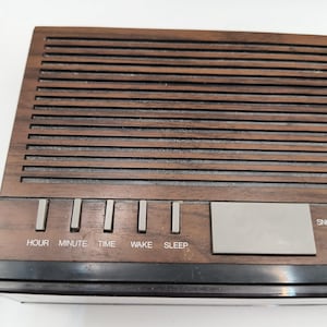 May include: A retro digital clock radio with a wood-effect finish. The clock has buttons marked "HOUR", "MINUTE", "TIME", "WAKE", and "SLEEP". A silver rectangular button is also visible. The clock has a black base.