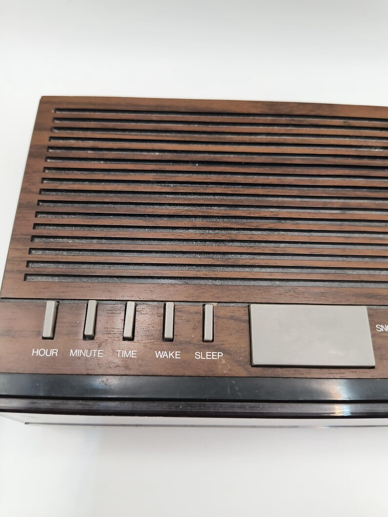 May include: A retro digital clock radio with a wood-effect finish. The clock has buttons marked "HOUR", "MINUTE", "TIME", "WAKE", and "SLEEP". A silver rectangular button is also visible. The clock has a black base.