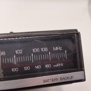 May include: Close-up of a black radio dial with white markings and text. The dial displays frequency numbers in MHz and kHz, with the text "BATTERY BACKUP" at the bottom. The dial is encased in a black frame.