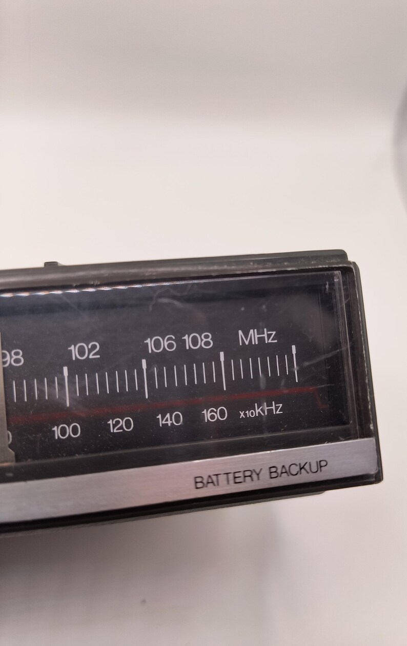 May include: Close-up of a black radio dial with white markings and text. The dial displays frequency numbers in MHz and kHz, with the text "BATTERY BACKUP" at the bottom. The dial is encased in a black frame.