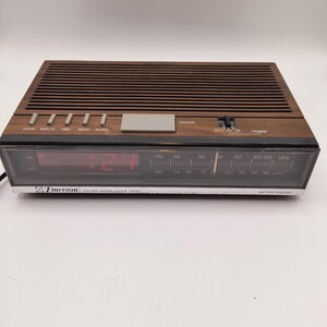 May include: A vintage Emerson AM/FM digital clock radio with a brown wood-effect finish. The clock displays red digital numbers and has buttons for hour, minute, time, wake, and sleep. It features a snooze button and an AM/FM radio dial.