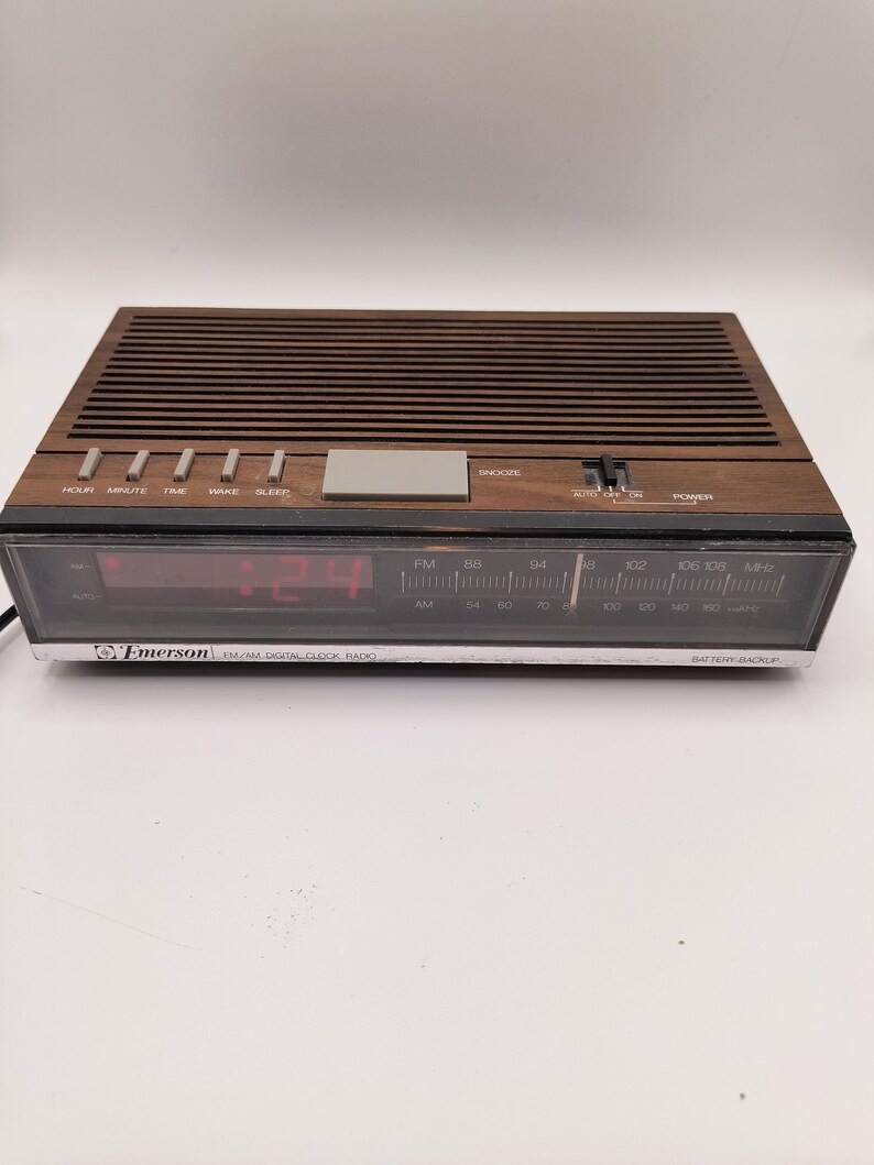 May include: A vintage Emerson AM/FM digital clock radio with a brown wood-effect finish. The clock displays red digital numbers and has buttons for hour, minute, time, wake, and sleep. It features a snooze button and an AM/FM radio dial.