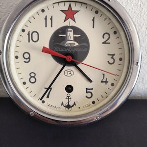 May include: A round wall clock with a silver metal frame and a cream-colored face. It features black Arabic numerals, a red star at the top, and a central graphic of a submarine with the text "Командирские". An anchor icon and "СДЕЛАНО В СССР" are printed at the bottom.