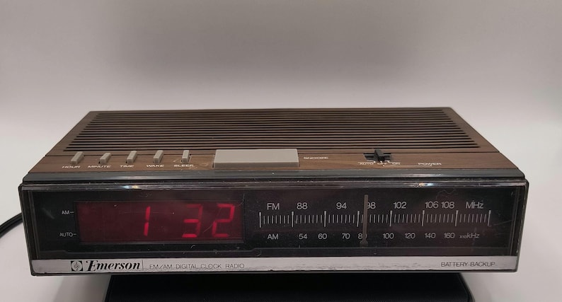 May include: A retro Emerson FM/AM digital clock radio with a brown wood-effect finish. The red digital display shows 1:32. Features include radio frequency dial, alarm, and battery backup.