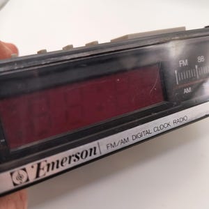 May include: Black Emerson FM/AM digital clock radio with a red digital display. The clock radio has a silver trim and grey buttons. The text "Emerson" and "FM/AM DIGITAL CLOCK RADIO" are visible on the device.