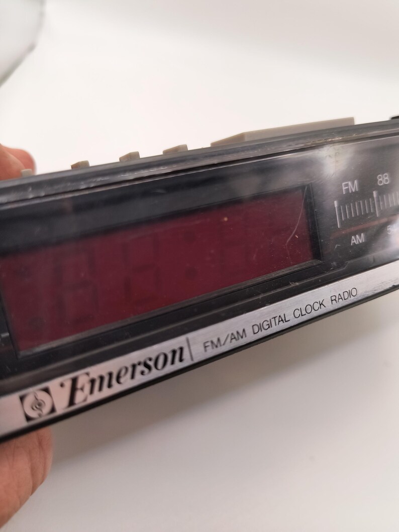 May include: Black Emerson FM/AM digital clock radio with a red digital display. The clock radio has a silver trim and grey buttons. The text "Emerson" and "FM/AM DIGITAL CLOCK RADIO" are visible on the device.