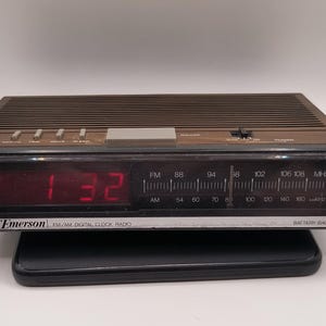 May include: A vintage Emerson FM/AM digital clock radio with a brown top and a black front panel. The red digital display shows the time as 1:32. Features include radio frequency dials and function buttons.