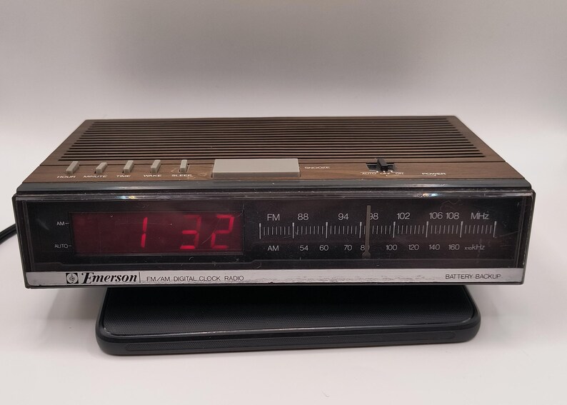 May include: A vintage Emerson FM/AM digital clock radio with a brown top and a black front panel. The red digital display shows the time as 1:32. Features include radio frequency dials and function buttons.