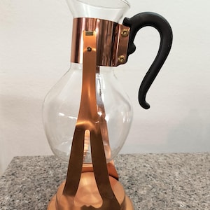 May include: A clear glass coffee maker with copper-colored metal accents and a black handle. The coffee maker has a round base and a bulbous glass body, ideal for brewing coffee. The design is vintage and elegant.