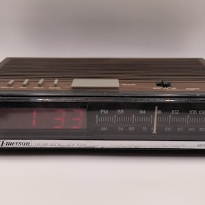 May include: A brown Emerson FM/AM digital clock radio displaying 1:33 in red. The radio features a tuning dial for FM and AM frequencies and a battery backup function.