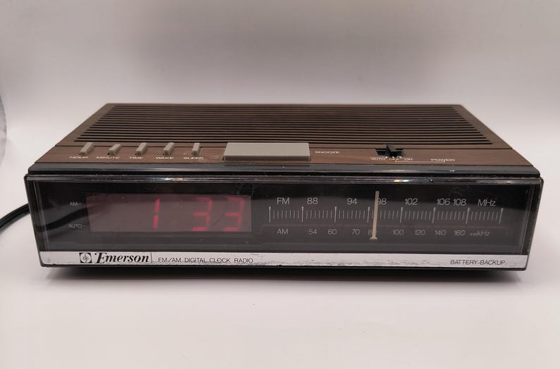 May include: A brown Emerson FM/AM digital clock radio displaying 1:33 in red. The radio features a tuning dial for FM and AM frequencies and a battery backup function.