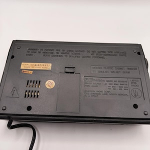 May include: A black Emerson radio, model RE05611B, with a cabinet finished to simulate walnut grain. The back of the radio has a serial number and warning labels. The radio has an AC power source.