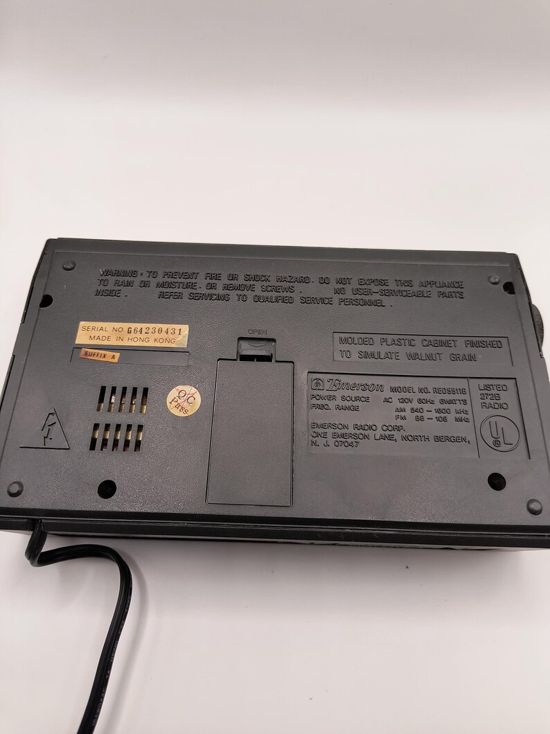 May include: A black Emerson radio, model RE05611B, with a cabinet finished to simulate walnut grain. The back of the radio has a serial number and warning labels. The radio has an AC power source.