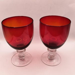 May include: Two red wine glasses with clear stems and bases. The bowls of the glasses are a deep red, transitioning to a lighter orange hue at the top. The stems are clear and feature a decorative design.