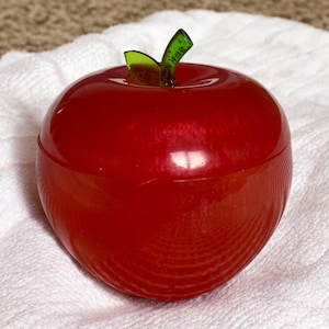 May include: A shiny red apple-shaped container with a green stem and leaf. The container is on a white, textured surface. The apple-shaped container is a decorative item.