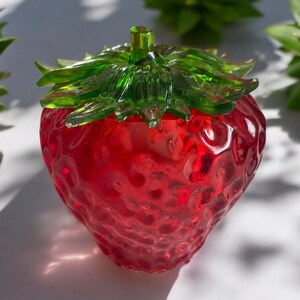 May include: A red, glass strawberry-shaped container with a green leafy top. The container has a textured surface and is likely a decorative item or a small trinket box. The image is well-lit, highlighting the details of the glass.