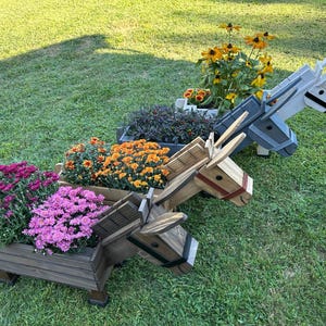 Könnte beinhalten: Drei hölzerne Esel-Pflanzgefäße gefüllt mit bunten Blumen. Die Pflanzgefäße sind in Braun-, Grau- und Weißtönen lackiert. Die Blumen umfassen rosa, orange und gelbe Blüten und schaffen eine lebendige Gartenausstellung. Die Pflanzgefäße stehen auf einem grünen Rasen.