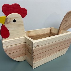 May include: A wooden chicken-shaped planter with a red comb and wattle, a yellow beak, and a natural wood finish. The planter has a rectangular compartment for holding plants or other items. The tail is made of wood. The planter is approximately 30 cm long.