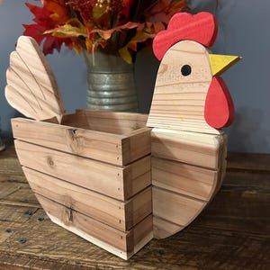 Könnte beinhalten: Eine holzgeschnitzte Box in Form eines Hahns mit einem Klappdeckel. Der Hahn hat einen roten Kamm und Kehllappen, einen gelben Schnabel und ein schwarzes Auge. Die Box ist aus hellem Holz gefertigt und für kleine Gegenstände konzipiert. Die Box steht auf einer Holzoberfläche.
