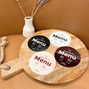 May include: Four circular menu signs with the text "Tap to View Our Menu" in white, black, and red. The signs are on a wooden cutting board. A small white vase with lavender sprigs is in the background.