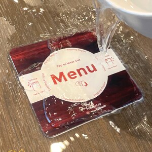 May include: A rectangular menu card with a dark red wood grain design is being splashed with water. The card features the word "Menu" in large red letters, with the text "Tap to View Our" above it. The card also has the text "Tapping Region" and "Tap on Back".