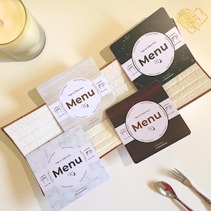 May include: Four square menu cards with different surface designs: marble, wood grain, and stone. Each card has a white circle with the word "Menu" and the text "Tap to View Our". A candle and silverware are in the background.