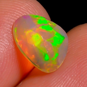 May include: A vibrant, oval-shaped opal gemstone. The opal displays a play of color with flashes of green, yellow, and orange hues. The gemstone is held between fingers, showcasing its polished surface and internal iridescence. The background is dark.