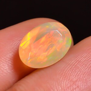 Puede incluir: Una piedra preciosa de ópalo de forma ovalada con una superficie facetada. El ópalo muestra un vibrante juego de colores, con destellos de naranja, amarillo y verde. La piedra preciosa se sostiene en una mano sobre un fondo oscuro.