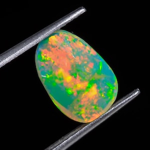 Puede incluir: Una vibrante piedra preciosa de ópalo, sujeta con pinzas plateadas, sobre un fondo negro. El ópalo muestra una mezcla de colores, incluyendo naranja, amarillo, verde y azul, con una superficie pulida y lisa. La gema tiene forma ovalada.