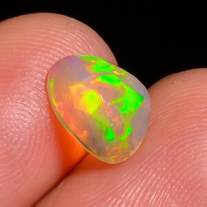 Puede incluir: Una vibrante piedra preciosa de ópalo con forma de pera. La piedra muestra una mezcla de colores, incluyendo tonos verdes brillantes, amarillos y naranjas, sobre una base blanca lechosa. El ópalo se sostiene entre dos dedos, mostrando su tamaño y brillo.