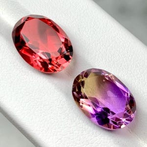 May include: Two oval-cut gemstones. One is a vibrant red-orange color, and the other is a gradient of yellow to purple. The gems are set against a white surface, showcasing their facets and brilliance. Jewelry.