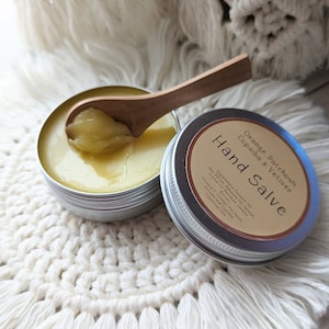 May include: A small silver tin with a wooden spoon scooping out a yellow hand salve. The tin has a label that reads "Orange Patchouli, Copaiba & Vetiver Hand Salve" and lists the ingredients.