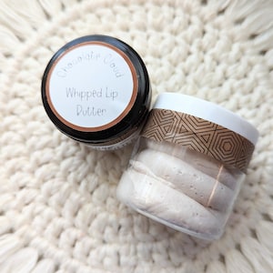 May include: Two small jars of lip butter on a white, textured surface. One jar is black with a white label that reads "Chocolate Cloud Whipped Lip Butter." The other jar is clear with a geometric patterned band and contains several white, oblong lip butter pieces.