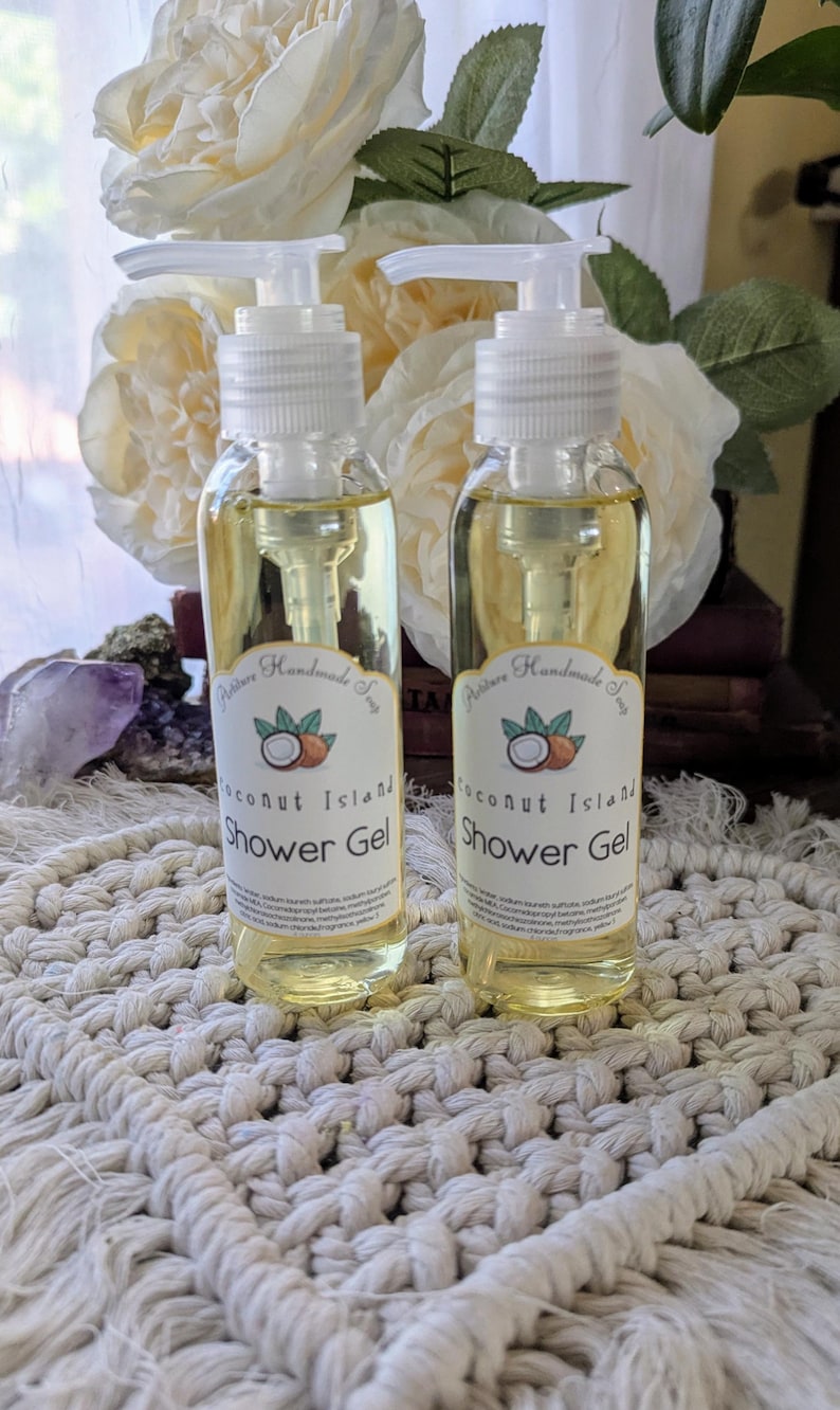 May include: Two clear bottles of Coconut Island shower gel with white pump tops. Each bottle has a label with a coconut illustration. The bottles sit on a white macrame surface, with white flowers in the background.
