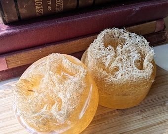 May include: Two round, amber-colored soap bars with a natural loofah texture. The bars are sitting on a wooden surface.