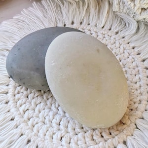 May include: Two smooth, oval-shaped soaps. One is a light gray color, and the other is off-white. They rest on a white macrame coaster with a fringed edge. The background includes more macrame and a hint of a pink flower.