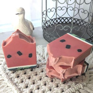 May include: Rectangular bars of soap designed to resemble watermelon slices, featuring a pink interior with embedded black seeds, a thin white rind, and a dark green outer edge. The soap bars have a textured top and are stacked in a group.