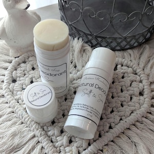 May include: Three white deodorant containers labelled "Salt & Sea" and "Natural Deodorant". One is a small jar, and two are stick deodorants. A white duck ornament and a decorative metal basket are also in the image.