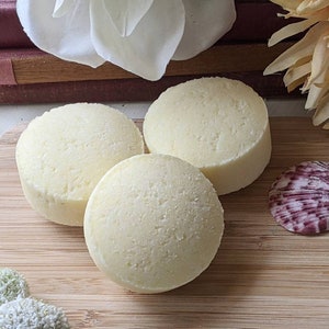 May include: Three white, round, solid shampoo bars on a wooden surface. The bars have a grainy texture.