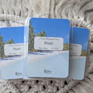 May include: Three white wax melt containers with a beach scene and blue sky. The text on the container reads "Artiture Handmade Soap", "Beach", "Wax Melts", and "2.75 OZ".