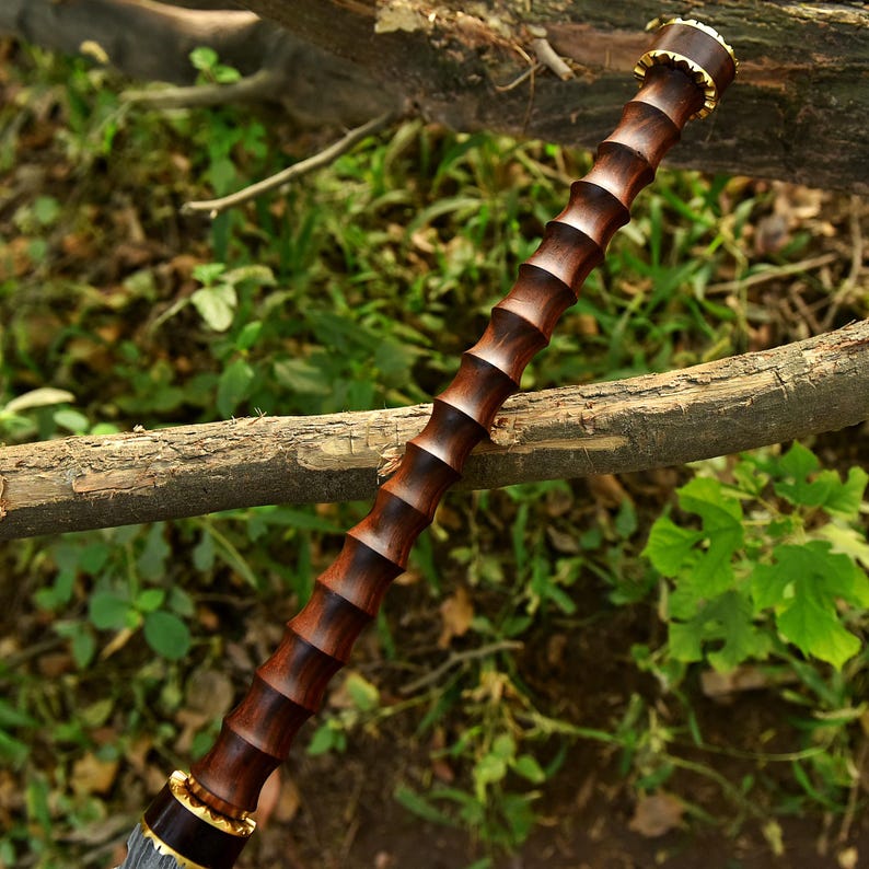 May include: A dark brown wooden walking stick with a ridged design. The top and bottom of the stick have gold-colored accents. The stick is resting on a tree branch with a green and brown background.