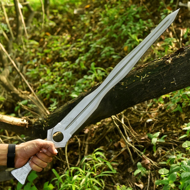May include: A silver-colored sword with a long, double-edged blade. The hilt features a circular opening and decorative engravings. The sword is held in a hand with a black watch. The background is a natural outdoor setting with green foliage.