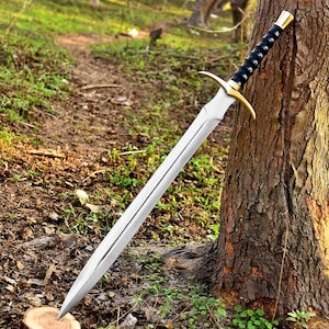 May include: A long, silver sword with a double-edged blade, gold crossguard, and black handle. The sword is leaning against a tree in a natural outdoor setting, with a focus on the blade's sharp point and the detailed hilt.